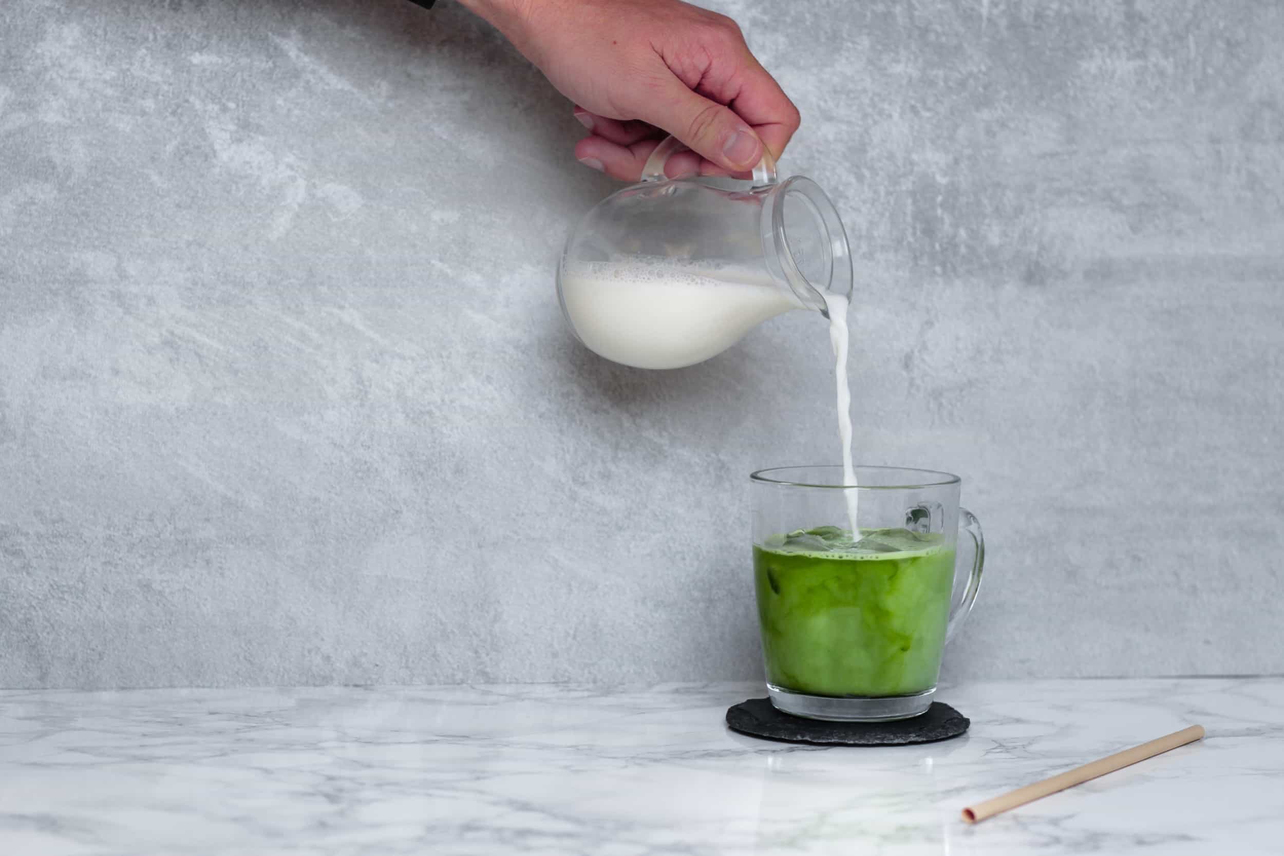 Laufende Zubereitung eines kalten Matcha-Latte. Man gießt die Milch in eine Tasse mit Matcha-Pulver.