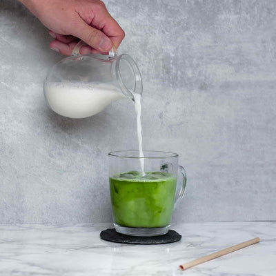 Zubereitung eines Matcha-Latte-Getränks mit traditionellem Bio-Matcha von matchadays. Man sieht eine Hand, die Milch in eine Tasse gießt.